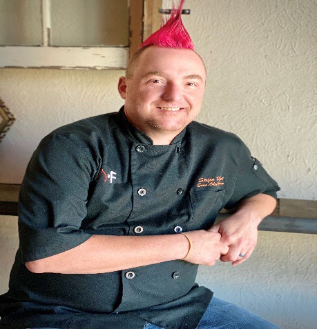 Chef with a bright pink mohawk and a black uniform smiling confidently.